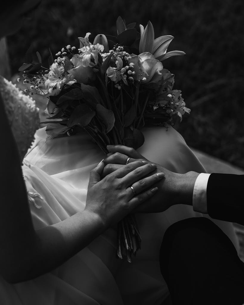 A touching black and white shot of a newlywed couple holding hands with a bouquet.